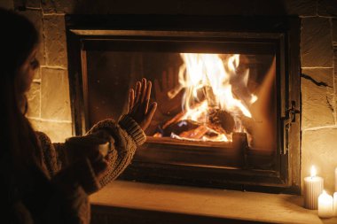 Kadın elleri çay bardağını tutuyor ve karanlık bir akşam odasında şöminenin önünde ısınıyor. Kapatın. Evde şömine ısıtması, elektrik kesintisi. Evde şömine başında atmosfer zamanı