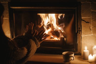 Woman hands warming up at cozy fireplace in dark evening room, close up. Fireplace heating in house, electricity blackout. Atmospheric time at fireside in home clipart