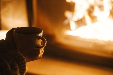 Karanlık bir akşam odasında bir fincan çay tutan ve sıcak bir şöminede elini ısıtan kadın, kapat. Evde şömine ısıtması, elektrik kesintisi. Evde şömine başında atmosfer zamanı