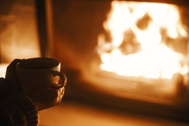 Karanlık bir akşam odasında bir fincan çay tutan ve sıcak bir şöminede elini ısıtan kadın, kapat. Evde şömine ısıtması, elektrik kesintisi. Evde şömine başında atmosfer zamanı