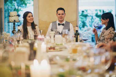 Beautiful wedding couple celebrating wedding on background of delicious festive table with food and drinks in restaurant. Stylish happy bride and groom with guests. Provence wedding reception