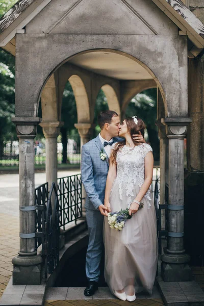 Stylish bride and groom kissing and embracing on background of old church. Romantic moment. Provence wedding. Beautiful sensual wedding couple gently hugging in european city