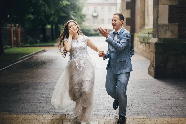 Stylish happy bride and groom running on background of old church in rainy street. Provence wedding. Beautiful emotional wedding couple smiling and having fun in rain in european city.