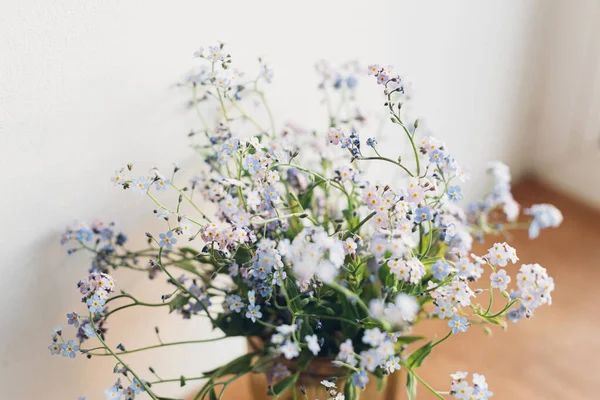 Beautiful little blue flowers in vase in warm sunlight on rustic wooden background. Delicate myosotis petals, forget me not spring flowers. Atmospheric evening moment. Simple countryside living