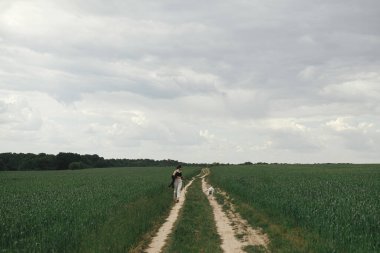Woman walking with cute white dog in wheat field. Stylish young female hiking and running with funny dog in summer countryside. Pet and travel. Danish spitz