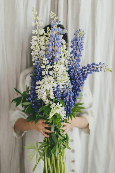 Stylish woman behind lupine bouquet in rustic room. Gathering and arranging summer wildflowers at home in countryside. Lupine flowers close up in hands of young female in linen dress