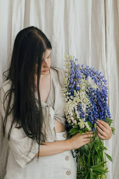 Young female in linen dress posing with lupine flowers. Gathering and arranging summer wildflowers at home in countryside. Stylish woman holding lupine bouquet in rustic room, close up