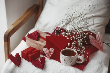 Happy valentines day! Cute stylish cup of tea with heart on cozy modern armchair with gifts, red envelope and white flowers. Valentine morning surprise for beloved