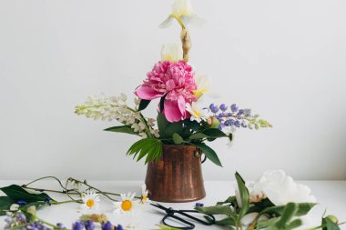 Modern summer flowers still life arrangement in vintage vase on white wood. Creative floral image. Stylish peony, lupin, iris and daisy composition and scissors on rustic table