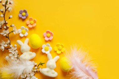 Happy Easter! Easter flat lay with eggs, flowers, bunny, feathers and blooming cherry on yellow background. Modern festive template with space for text. Greeting card or banner