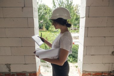 Tabletli genç bayan mimar yeni modern evin planlarını kontrol ediyor. İnşaat alanındaki dijital planlara bakan şapkalı şık, mutlu kadın mühendis.