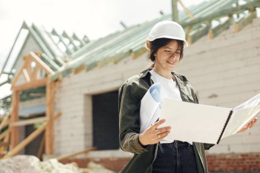 Genç bayan mühendis ya da inşaat işçisi yeni modern bir ev inşa etmeye karşı gülümsüyor. İnşaat alanında planları ve planları olan şık, mutlu bir mimar.