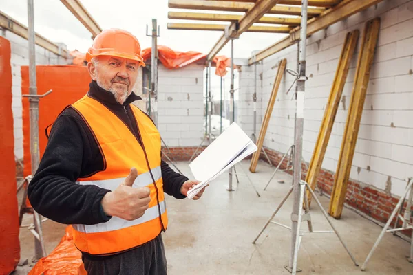 Senior man engineer or construction worker in hardhat approving building new modern house with blueprints. Portrait of male architect or contractor holding plans at construction site