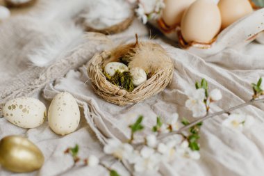 Happy Easter! Stylish easter eggs and blooming spring flowers on rustic table. Rustic easter still life. Modern eggs in nest and cherry blossoms on linen fabric