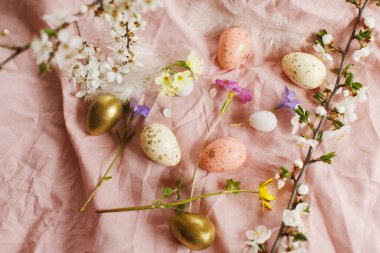 Stylish easter eggs and blooming spring flowers on pink linen fabric. Happy Easter! Modern eggs, feathers and cherry blossoms. Rustic easter still life