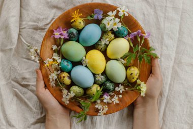 Stylish easter eggs and blooming spring flowers in wooden bowl in woman hands. Happy Easter! Rustic easter flat lay. Natural painted eggs and blossoms on rural table