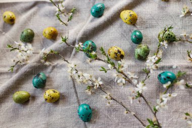 Happy Easter! Stylish easter eggs and blooming cherry branch on rustic table. Rustic easter flat lay. Natural painted quail eggs and spring blossom on linen fabric
