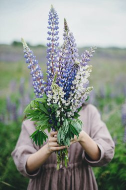 Close up of woman in rustic dress holding lupine bouquet in meadow. Cottagecore aesthetics. Young female in linen dress holding wildflowers in atmospheric summer countryside, rural slow life
