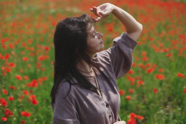 Stylish sensual woman in rustic dress relaxing in red poppy field. Cottagecore aesthetics. Young female in linen dress enjoying summer among wildflowers meadow in sunny countryside, slow life