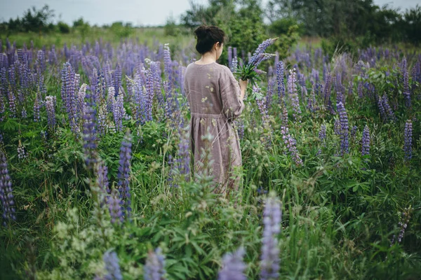 穿着乡村服装的时髦女人在草地上采摘羽扇豆 大气形象 土工布美学 穿着亚麻布衣服的年轻女子在夏天的乡间采花 农村的生活缓慢 — 图库照片