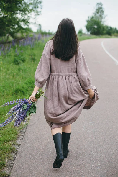 夏天的乡间 穿着乡村风情服装 头戴紫丁香花束的时髦女人在散步 土工布美学 穿着亚麻布衣服的年轻女子 雨后抱着野花在乡间路上 生活节奏缓慢 — 图库照片