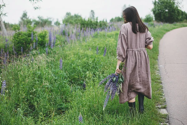 土工布美学 夏天的乡间 穿着乡村风情服装 头戴紫丁香花束的时髦女人在散步 穿着亚麻布衣服的年轻女子 雨后抱着野花在乡间路上 生活节奏缓慢 — 图库照片