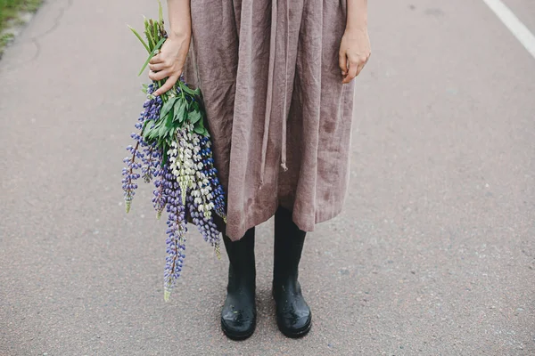 在乡村的夏天 穿着粗俗衣服和长统靴的女人手里拿着淡紫色的花束 土工布美学 乡村道路上的雨后 年轻女子手持野花 — 图库照片