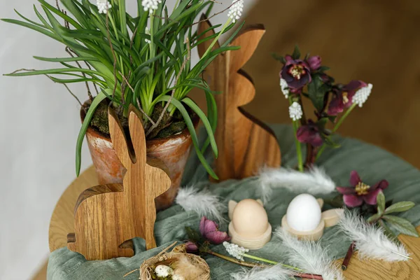 Easter still life. Stylish wooden bunny, spring flowers and natural eggs on rustic table in room. Happy Easter! Festive arrangement and decor in farmhouse