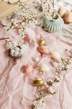 Stylish easter eggs and blooming spring flowers on pink linen fabric. Happy Easter! Modern eggs in nest, feathers and cherry blossoms. Rustic easter still life