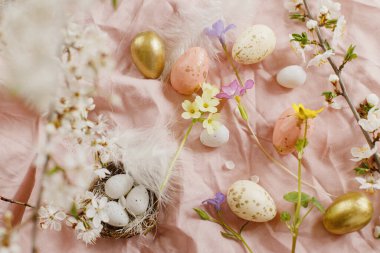 Happy Easter! Stylish easter eggs and blooming spring flowers on pink linen fabric. Modern eggs in nest, feathers and cherry blossoms. Rustic easter still life
