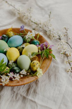 Stylish easter eggs and blooming spring flowers in wooden bowl on linen fabric. Happy Easter! Rustic easter still life. Natural painted eggs and blossoms on rural table