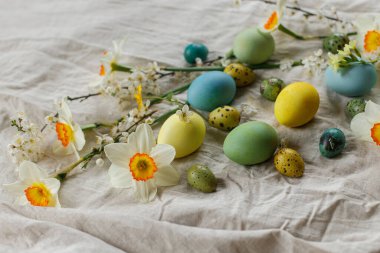 Happy Easter! Stylish easter eggs and blooming spring flowers on rustic table. Rustic easter still life. Natural painted eggs and daffodils bloom on linen fabric