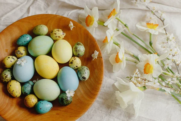 Happy Easter!  Rustic easter flat lay. Stylish easter eggs and blooming daffodils flowers in wooden bowl on rustic table. Natural painted eggs and blossoms on linen fabric