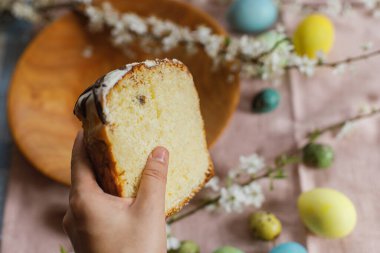Hand holding half of baked easter cake on background of natural dyed easter eggs with spring flowers on wooden plate on rustic table.  Traditional Easter food. Happy Easter!