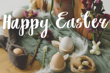 Tahta tavşan kulakları, doğal paskalya yumurtaları, bahar çiçekleri, kırsal masadaki tüyler üzerine Mutlu Paskalya yazıları. Modern Paskalya tebrik kartı. El yazısıyla yazılmış harfler