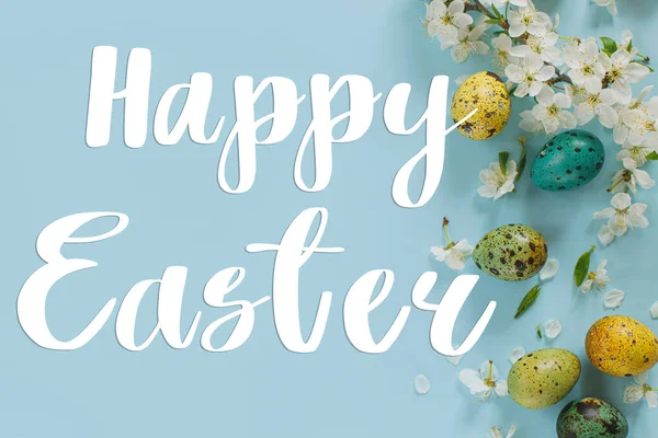 Happy Easter greeting card. Happy Easter text on Easter stylish eggs and blooming spring flowers on blue background flat lay. Modern Greeting card. Handwritten sign