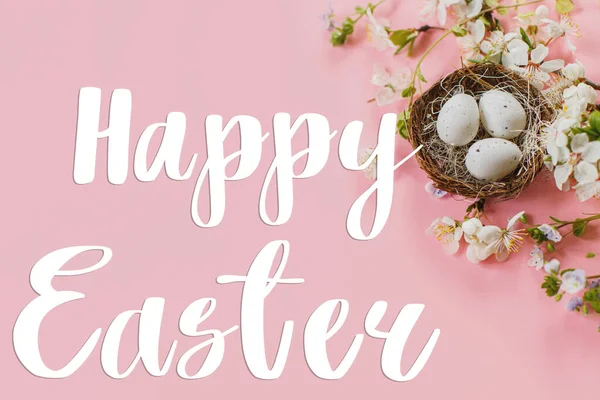 Happy Easter greeting card. Happy Easter text on Easter eggs in nest and cherry blossoms flat lay on pink background. Modern Greeting card. Handwritten sign
