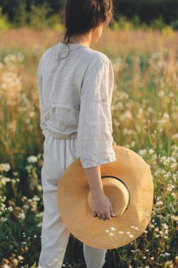 Sıcak günbatımı ışığında kır çiçeklerinin arasında yürüyen hasır şapkalı şık bohem kadın. Yaz tatili ve seyahat. Kırsal keten kumaşlı genç bir kadın yaz otlaklarında dinleniyor. Atmosferik an