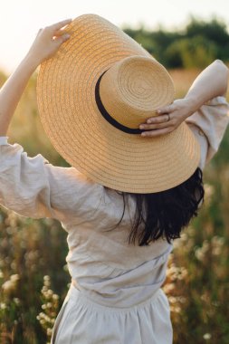 Gün batımı ışığında kır çiçeklerinin arasında poz veren hasır şapkalı şık bohem kadın. Yaz zevki ve sükunet. Kırsal keten kumaş içinde kaygısız genç bir kadın yaz çayırında dinleniyor.