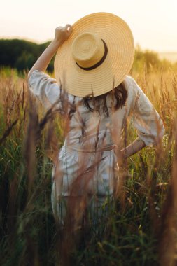Hasır şapkalı şık boho kadın gün batımı ışığında yabani otların arasında poz veriyor. Yaz zevki ve sükunet. Kırsal keten kumaş içinde kaygısız genç bir kadın yaz çayırında dinleniyor.