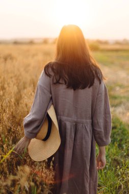 Hasır şapkalı şık bir kadın güneş ışığında yulaf tarlasında yürüyor. Atmosferik sükunet anı. Keten elbiseli genç bir kadın yaz akşamları dinleniyor, kırsal kesimde sakin bir hayat sürüyor.