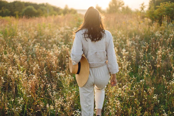 在温暖的夕阳西下 戴着草帽的时髦女人在野花间漫步 大气瞬间 夏天的快乐和旅行 穿着乡村亚麻布的年轻女子在夏日的草地上悠闲自在 — 图库照片