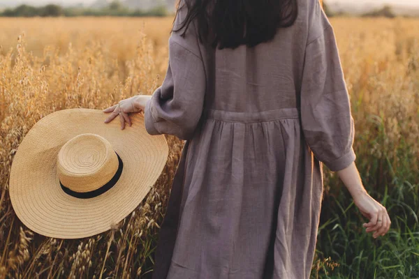 头戴草帽的时髦女人在夕阳西下走在燕麦田边 后视镜 大气宁静的时刻 穿着亚麻布衣服的年轻女子在夏夜的乡间悠闲 乡村的悠闲生活 — 图库照片
