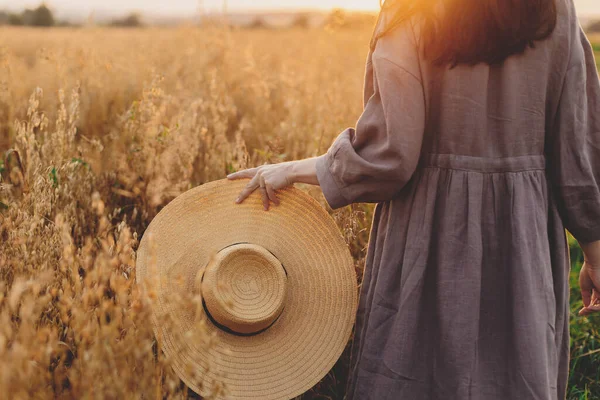 日没の光の中でオートフィールドを歩く麦藁帽子を持つスタイリッシュな女性 クロップドビュー 落ち着いた雰囲気の瞬間 若いです女性で素朴なリネンドレスで夜夏の田舎でリラックス — ストック写真