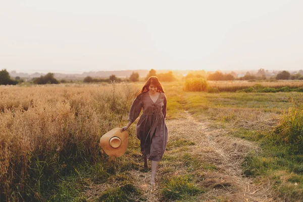 日没の光の中でオートフィールドで走る麦藁帽子を持つスタイリッシュな女性 幸せな瞬間だ 若いです女性で素朴なリネンのドレスでリラックスした夜夏の田舎 田舎生活 — ストック写真