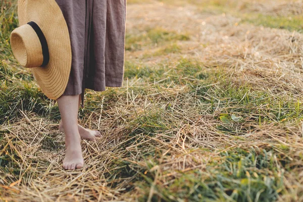 头戴帽子的女人赤脚走在落日下的稻草地里 关门了 农村缓慢的生活 大气宁静的时刻 夏乡的年轻女子穿着淡雅的亚麻布衣服 — 图库照片