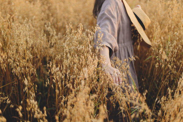 Stylish woman with straw hat holding oat stems in evening light. Rural slow life. Young female in rustic linen dress standing in harvest field in summer countryside. Atmospheric tranquil moment