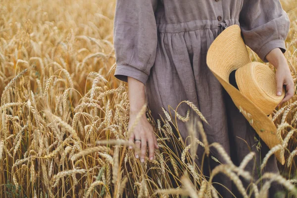 麦藁帽子をかぶった女性が畑で茎を保持します 落ち着いた雰囲気の瞬間 田舎の夏の夜に熟した小麦の耳に触れる素朴なリネンのドレスの女性 農村のスローライフ — ストック写真