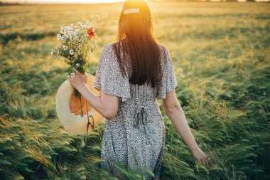 Gün batımında arpa tarlasında duran kır çiçekleriyle güzel bir kadın. Şık bir kadın yaz kırsalında dinleniyor ve çiçek topluyor. Atmosferik sükunet anı