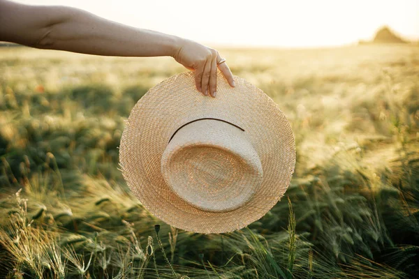 Straw hat in woman hand in sunset light on background of barley field. Stylish female enjoying evening summer countryside. Atmospheric tranquil moment, rustic slow life